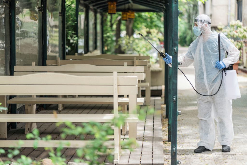 Sanitation worker in protective suit disinfecting benches and tables outdoors